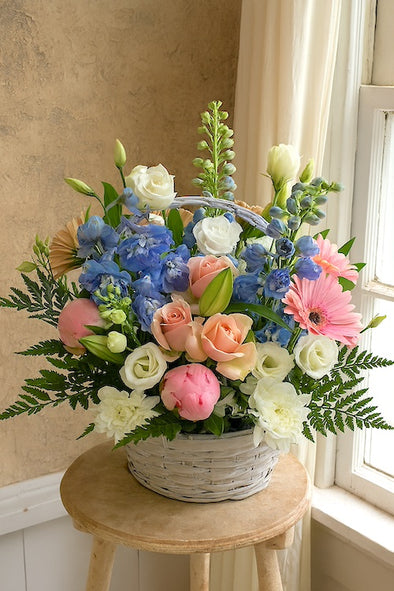 Elegant sympathy flower basket with soft pastel blooms, including blue delphinium, pale roses, lisianthus, peonies, and greenery in a wicker basket